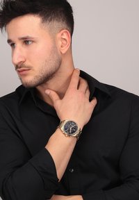 Guld-tone ur med en mørkeblå urskive, kronograffunktioner og et metalarmbånd. Model poserer med hånden på halsen iført en sort skjorte med knapper.