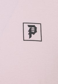 Camiseta de algodón de color rosa claro con un logo cuadrado negro que presenta una letra "P" estilizada en el centro. Textura suave y diseño ajustado.