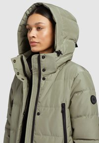 Jachetă puffer verde măsliniu cu glugă spațioasă, buzunare laterale cu fermoar, accesorii negre și un patch logo rotund pe mâneca stângă.