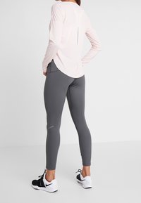 Lichtroze langemouwen top met afgeronde onderkant en splitten aan de achterkant, gedragen met slim-fit grijze leggings en zwart-witte sportschoenen.
