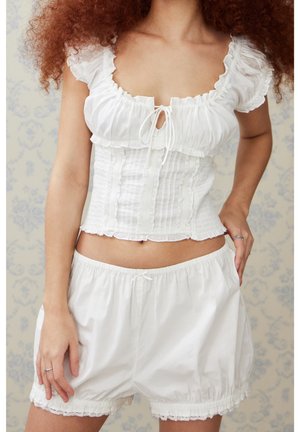 Woman wearing white ruffled crop top with puff sleeves and loose shorts standing against floral wallpaper background.