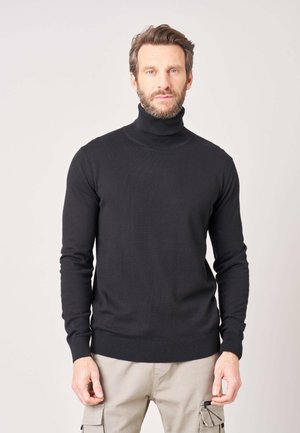 Man met kort haar en baard, gekleed in een zwarte coltrui en beige cargobroek, staand tegen een neutrale achtergrond.