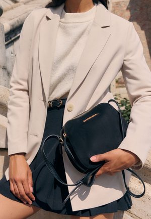 Kvinde iført cream blazer og sweater, navy plisseret nederdel, holder sort crossbody taske med gyldent lynlås og logo.