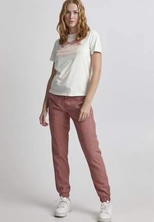 Cream t-shirt with a watercolor-style graphic in pink, paired with pink elastic cuff pants and white sneakers on a neutral background.