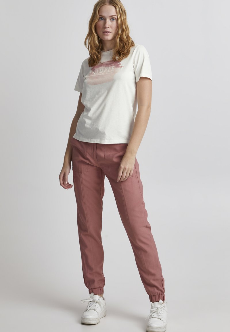 Crème t-shirt met een grafiek in aquarelstijl in het roze, gecombineerd met roze elastische broekspijpen en witte sneakers op een neutrale achtergrond.