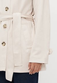 Beige button-up coat with a belted waist, featuring a soft texture, two side pockets, and eco-friendly buttons with a marbled pattern.