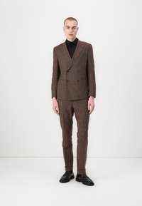Costum dublu cu model houndstooth maro, material texturat, croială slim, asortat cu o cămașă neagră și pantofi masivi negri. Fundal minimalist.