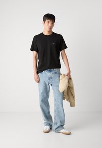 Black cotton t-shirt with short sleeves, blue distressed loose-fit jeans, and beige bomber jacket in hand. White sneakers with gum soles.