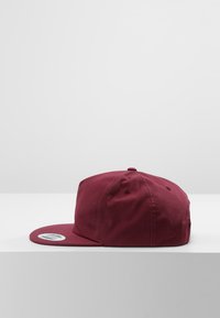 Bordó baseball sapka, szilárd szövetből készült, lapos karimájú és szellőzőlyukakkal rendelkezik, egy világosszürke felületen van bemutatva.