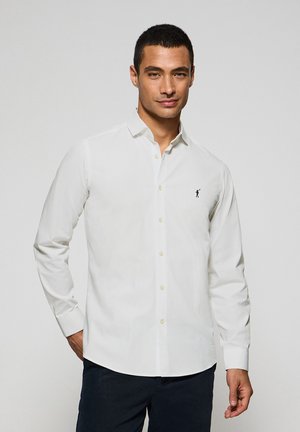 Camisa blanca de manga larga con botones y cuello clásico, que cuenta con un pequeño logo negro bordado en el pecho y una textura suave y lisa.