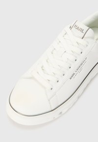 Sneaker bianca in pelle con lacci piatti, dotata di suola testurizzata, dettagli metallici e linguetta con marchio. Finitura liscia e design minimalista.