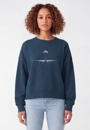 Jonge vrouw met krullend haar draagt een donkerblauwe trui met een kleine astronaut onder een gloeiende maan.