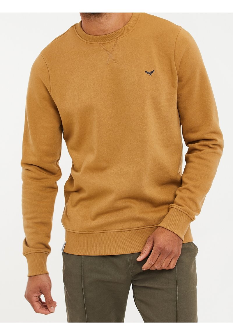 Homme portant un sweat-shirt à col rond couleur moutarde et un pantalon vert olive, debout avec une main proche de sa taille.