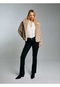 Blonda sieviete melna jostas, melnu pieguļošu biksu un melnu papēžu zābaku, valkājot bēša mākslīgās kažokādas jaku virs baltas blūzes, stāv pelēka fona priekšā.