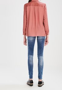 Růžová halenka s krajkovými detaily na ramenou, dlouhými rukávy a zadním skladem, spárována se slabě modrými skinny džínami a černými botami.