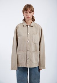 Chaqueta beige de gran tamaño con cuello, botones plateados y dos bolsillos frontales, hecha de un material similar al algodón, combinada con pantalones de mezclilla azules.