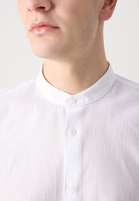Bílá, strukturovaná košile s límečkem a třemi knoflíky na přední straně. Hladká látka s jemným vzorem mřížky.