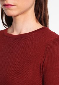 Nahaufnahme eines gerippten, burgunderfarbigen Textils im Rundhalsdesign, mit glatter Textur und enganliegender Silhouette.