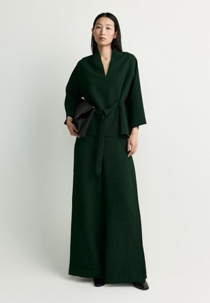 Woman wearing a dark green wide-leg jumpsuit with a belted waist, holding a large black clutch, standing against a plain white background.