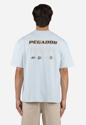 Homme aux cheveux foncés vu de dos, portant un t-shirt bleu clair de la marque Pegador et un pantalon beige sur fond uni.