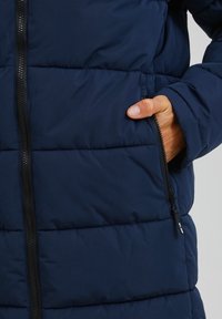 Marineblaue gepolsterte Jacke mit Reißverschluss und seitlichen Taschen, mit gesteppter Verzierung und glatter Textur. Reißverschlussakzente an den Taschen.