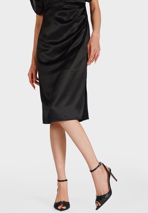 Donna che indossa una gonna nera di raso al ginocchio con uno spacco, posando con sandali neri con tacco alto a punta aperta e cinturini alla caviglia.