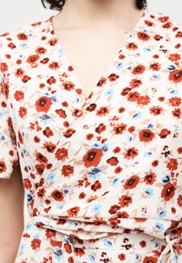 Κοντινό πλάνο λευκού φόρεματος τύπου wrap με κόκκινo, μπλε και καφέ λουλουδάτο μοτίβο, που φοριέται από ένα άτομο με κοντά σκούρα μαλλιά.