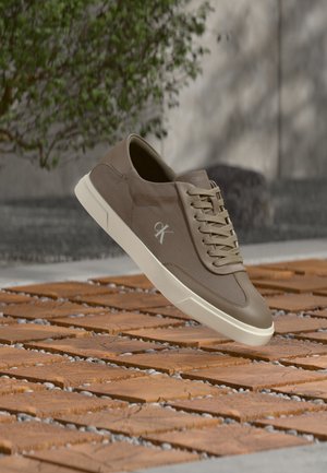 Baskets basses marron avec semelle blanche et logo 'ck' flottant au-dessus d'un sol carrelé en bois avec de la verdure et un mur en béton en arrière-plan.