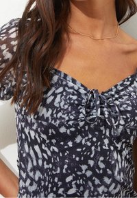 Mustapainokseton paita, jossa on neliönmuotoinen kaula-aukko, keskellä on rypytetty yksityiskohta ja nauhakoriste. Pehmeä kangas.