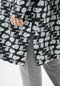 Schwarzes Hemd mit weißem Cartoon-Druck, das einen Charakter zeigt. Aus glattem Stoff, mit Knopfverschluss und einem seitlichen Schlitz. Graue Hose sichtbar.