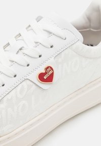 Zapatilla blanca con patrón en relieve "LOVE MOSCHINO", cordones blancos planos y una etiqueta de acento en forma de corazón rojo en el lateral. Suela de goma.