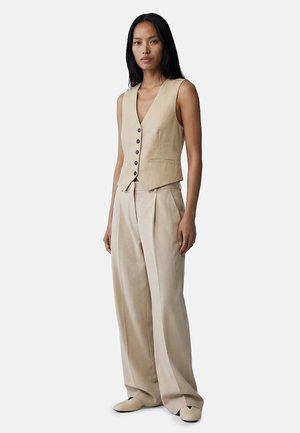 Kvinde med langt sort hår iført beige ærmeløs vest med knapper, vidde bukser og beige sko, stående mod en ensfarvet baggrund.