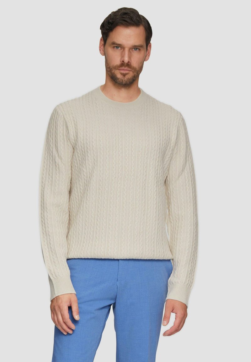 Helle beige Strickpullover mit Rundhalsausschnitt und gerippten Bündchen, kombiniert mit hellblauen Hosen. Texturiertes Muster über den Stoff.