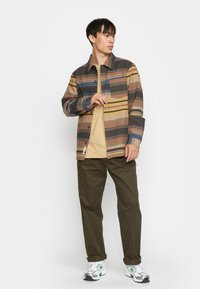 Gestreepte jas in neutrale tonen, met blauwe, gele en bruine lijnen. Draagt over een beige shirt, gecombineerd met olijfgroene broek en sneakers.