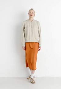 Lys beige bomberjakke med glidelås, kombinert med en oransje sateng midi-skirt og nøytrale sneakers. Glatte teksturer, uformell passform.