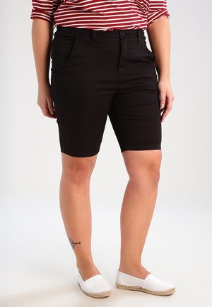 Person trägt schwarze knielange Shorts, rot-weiß gestreiftes Oberteil, weiße Slip-On-Schuhe und hat ein Tattoo am linken Knöchel, steht auf einem weißen Hintergrund.