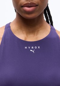 Κοντινό πλάνο ενός ατόμου που φοράει μωβ αθλητικό τοπ με τα λογότυπα "HYROX" και Puma στο στήθος και με ορατά κοτσίδια στα μαλλιά.