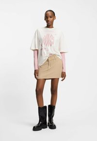 Hvid grafisk t-shirt lagdelt over en lyserød langærmet top, kombineret med en beige mininederdel og sorte knæhøje støvler.