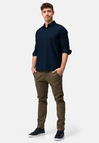 Marineblå skjorte med lange ærmer og knapper, khaki slim-fit bukser og mørke sneakers, poseret mod en hvid baggrund. Klassisk og afslappet styling.