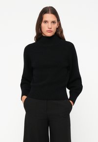 Schwarzer Strickrollkragenpullover mit geripptem Muster, figurbetontem Schnitt und langen Ärmeln. Kombiniert mit hochgeschnittenen schwarzen Hosen.