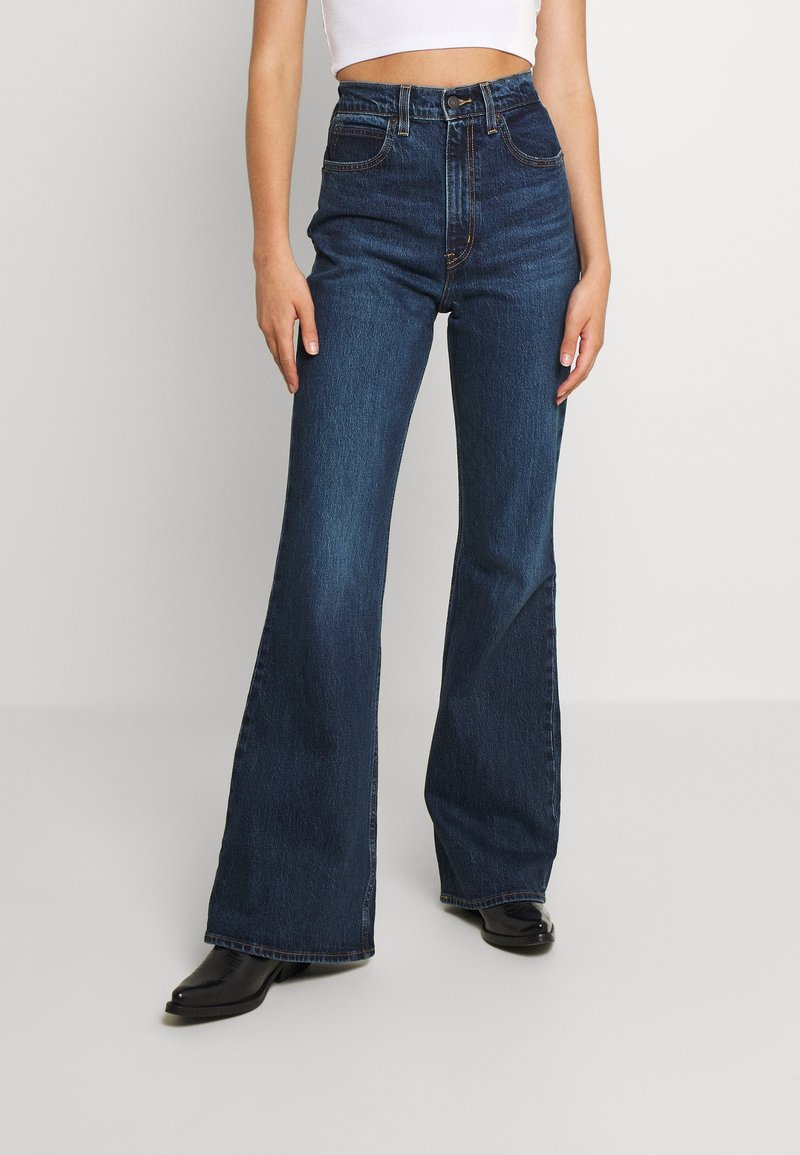 Femme portant un jean flare bleu foncé taille haute, un haut court blanc et des chaussures noires à bouts pointus, debout devant un fond blanc.