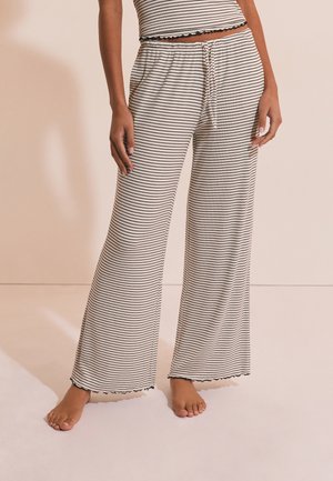 Femme portant un pantalon beige à jambes larges avec de fines rayures horizontales noires et un haut court assorti, debout pieds nus sur un fond beige.