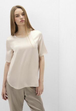 Giovane donna con capelli lunghi e lisci che indossa una camicetta beige larga a maniche corte e pantaloni marrone chiaro abbinati, in piedi contro un muro bianco semplice.