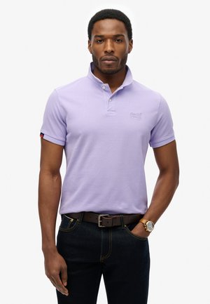 Camisa polo de manga corta en lavanda con cuello, que presenta un logo en el pecho y un acento rojo en la manga. Combinada con jeans oscuros y un cinturón marrón.