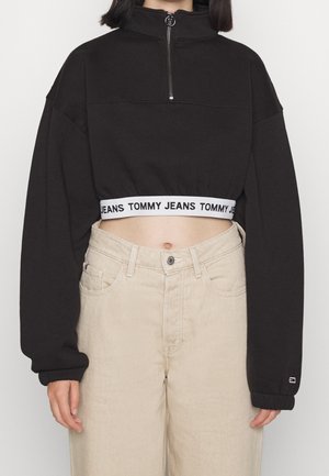 Musta lyhyt pusero, jossa vetoketju edessä ja Tommy Jeans -merkitty vyötärökaistale, puettuna korkeavyötäröisten beigejen housujen kanssa mallin vartalolla.