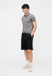 Grå polo-shirt med sort logo, sorte shorts med en hvid sideliste og beige sneakers. Model poserer med hænderne i lommerne.