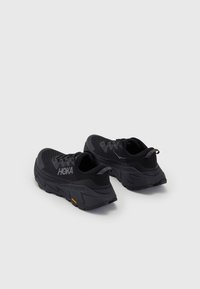 Mustat urheilukengät verkkokankaasta valmistetulla päällisellä, pehmeällä kauluksella, tekstuurilla varustetulla pohjalla ja huomattavalla HOKA-logolla. Ominaisuuksiin kuuluu paksu pohja, jossa on oranssi korostus.