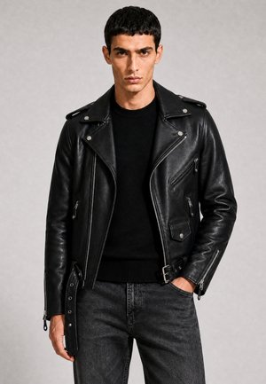 Mann trägt eine schwarze Leder-Bikerjacke über einem schwarzen Pullover und dunkelgrauen Jeans, steht mit einer Hand in der Tasche vor einem einfarbigen Hintergrund.
