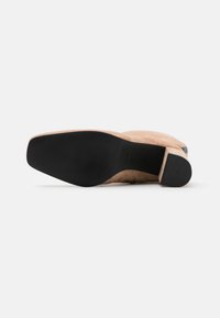 Chaussure à talons hauts en suede nude avec une semelle en caoutchouc noir, présentant une finition lisse, un bout rond et un accent de talon proéminent.