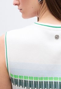 Κοντινό πλάνο στον ώμο και το λαιμό μιας γυναίκας που φοράει λευκό αμάνικο τοπ με πράσινες και μπλε ρίγες, και ένα μικρό στρογγυλό ασημένιο τσιμπιδάκι κοντά στον γιακά.